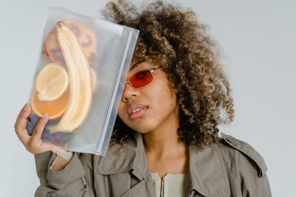 Young woman with curly hair holds a ziplock bag containing various fruits, indoors.